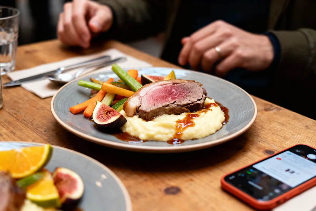 Assiette de viande saignante, purée, légumes variés, figues, sur table en bois, téléphone et verre à proximité.