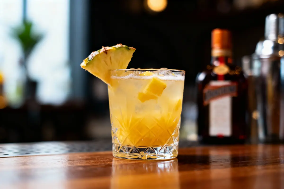 Cocktail jaune avec morceaux d'ananas, verre en cristal, fond flou, bouteille en arrière-plan.