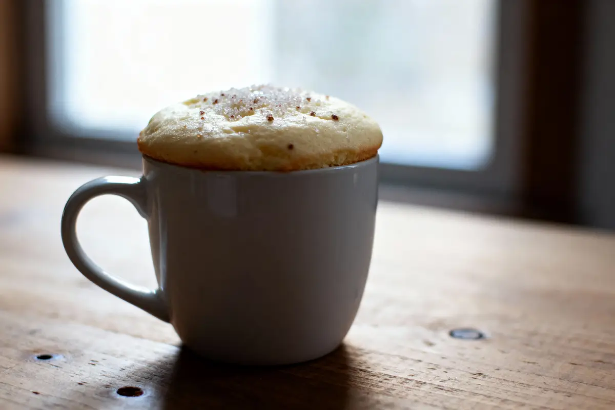 Tasse blanche contenant un gâteau soufflé, posée sur une table en bois devant une fenêtre floue.