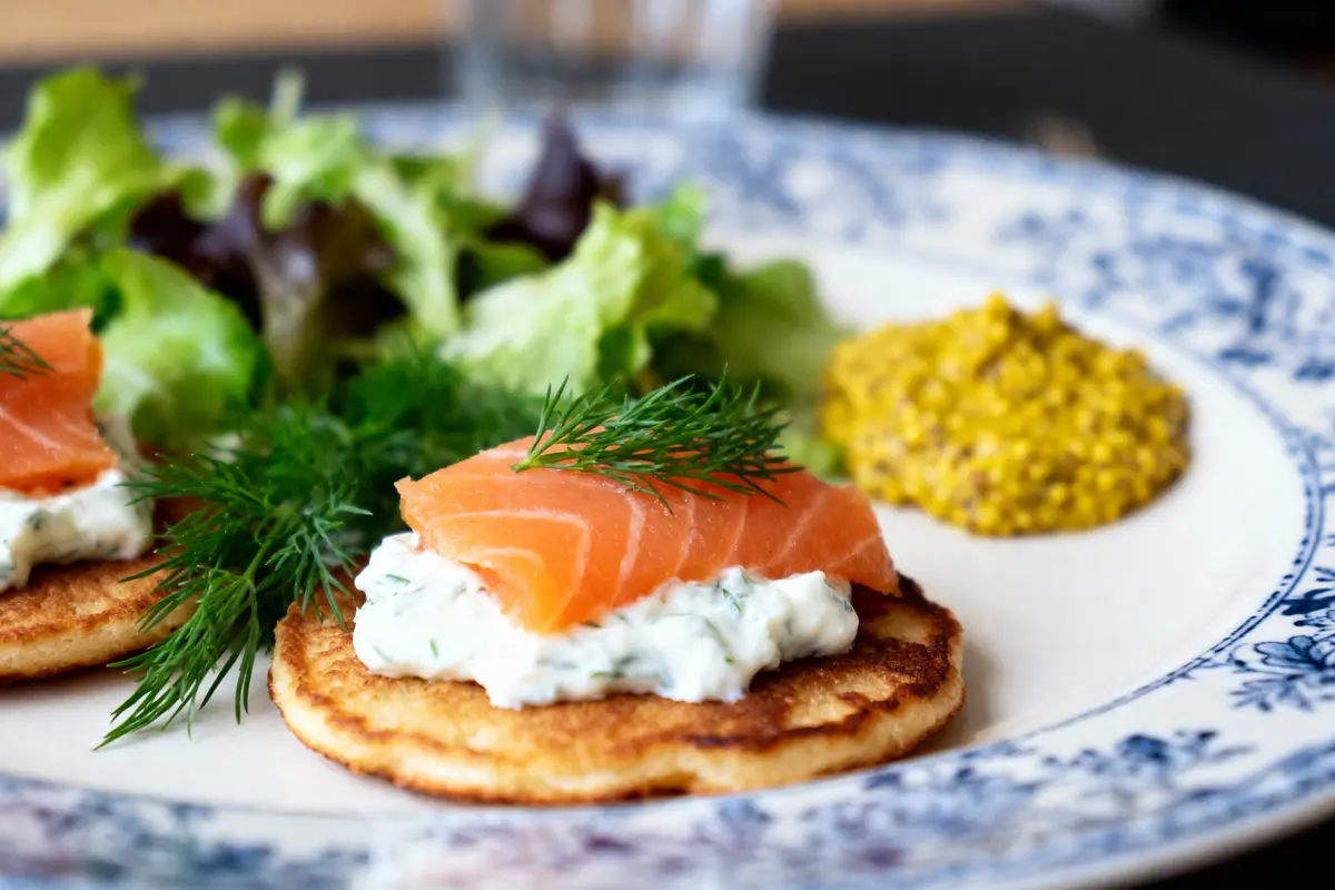 pexels-michal-pech-213601-757318 (1) Blini avec crème, saumon, aneth, servi avec salade verte et moutarde sur assiette blanche décorée.