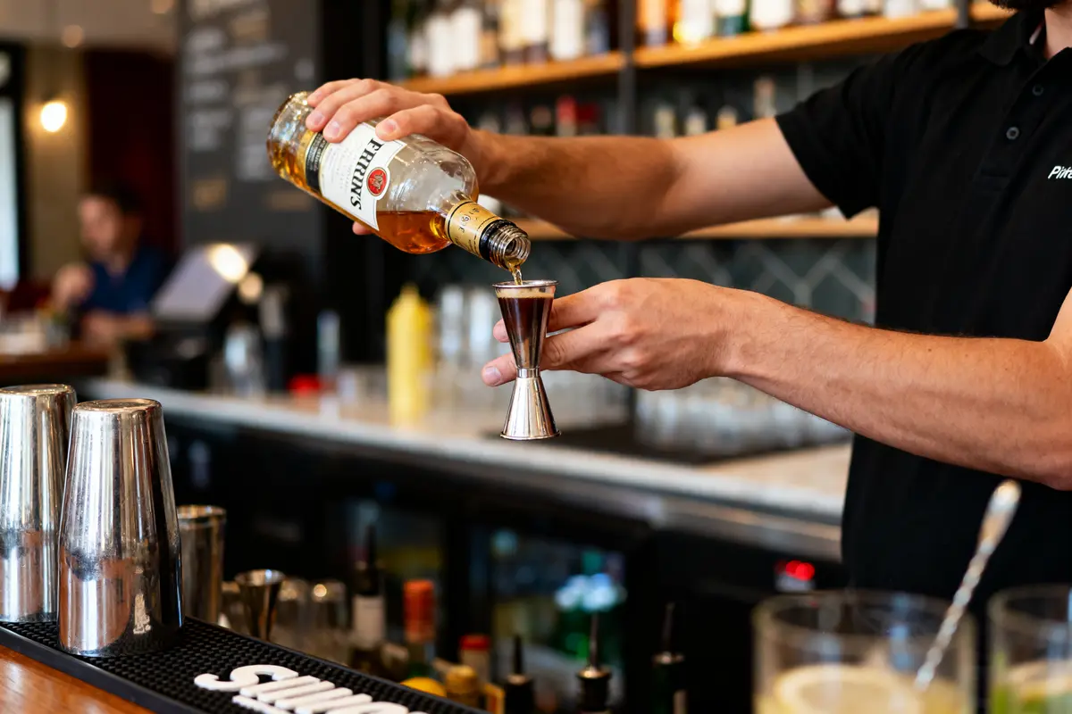 Serveur versant alcool dans jigger en métal, bar en arrière-plan avec bouteilles et équipements de mixologie.