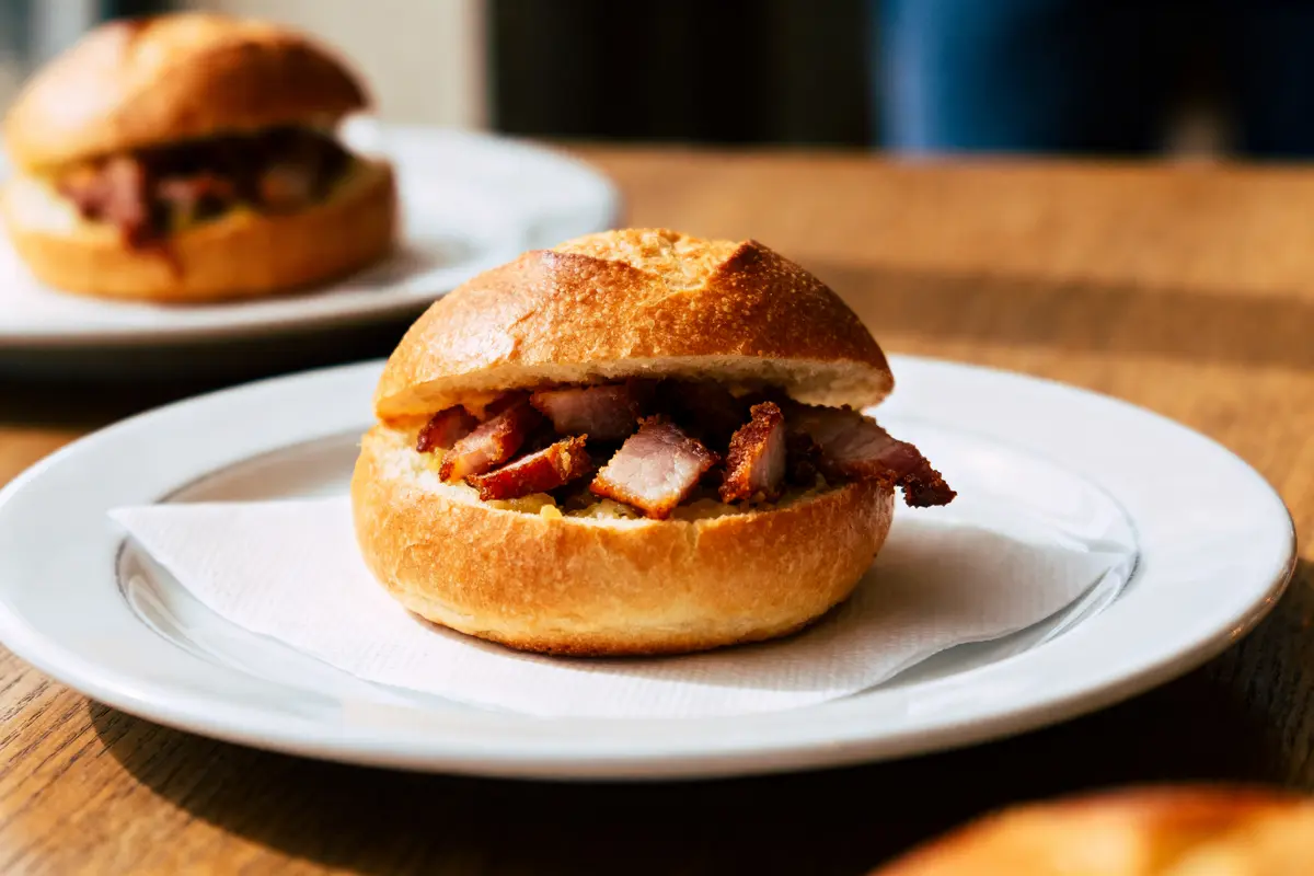 pexels-michal-pech-213601-757318 (1) Sandwich au porc croustillant dans un petit pain, sur une assiette blanche, servie sur table en bois.