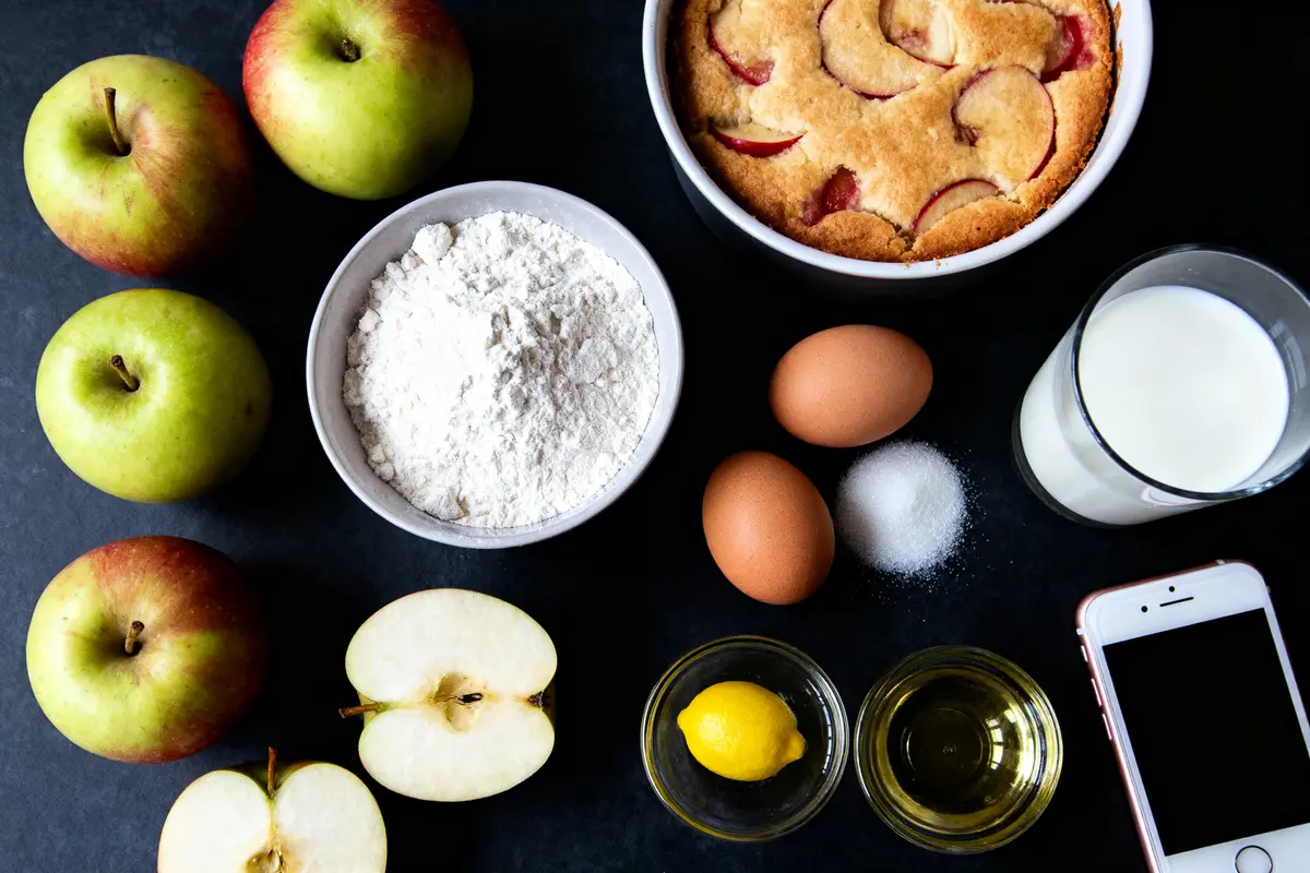Ingrédients pour gâteau aux pommes : pommes, farine, œufs, lait, sucre, huile, citron, téléphone, sur table noire.