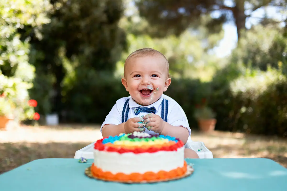 Bébé souriant avec gâteau d'anniversaire arc-en-ciel en extérieur, vêtements élégants, ambiance joyeuse.