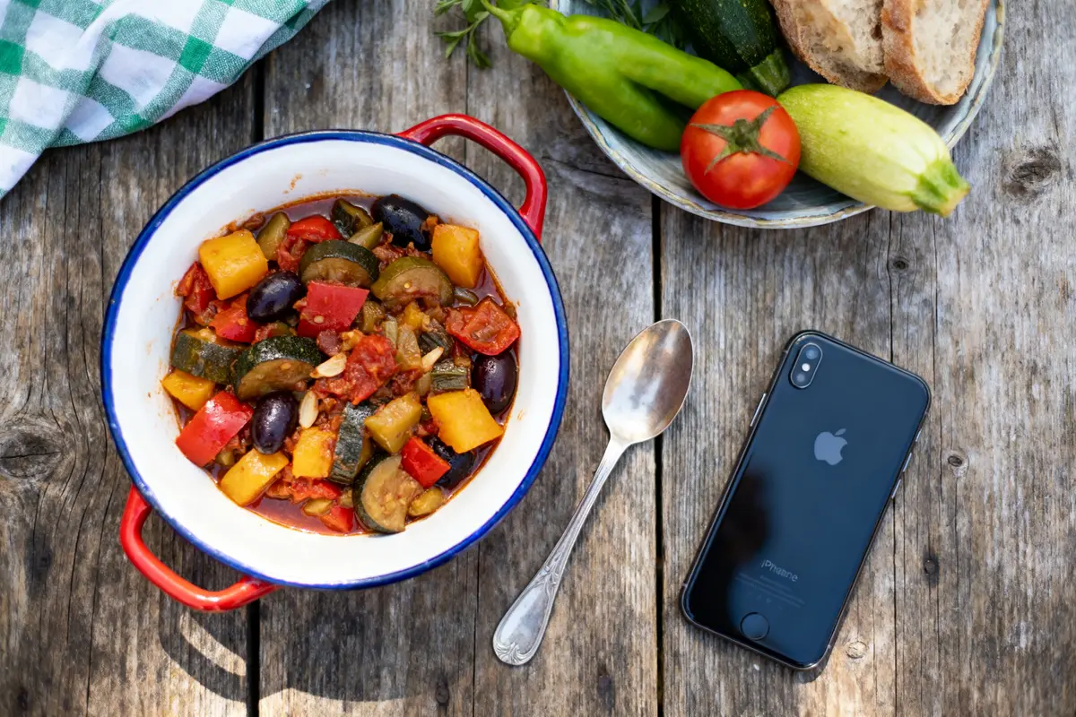pexels-michal-pech-213601-757318 (1) Bowl of ratatouille on wooden table with vegetables, bread, smartphone, and spoon beside it.