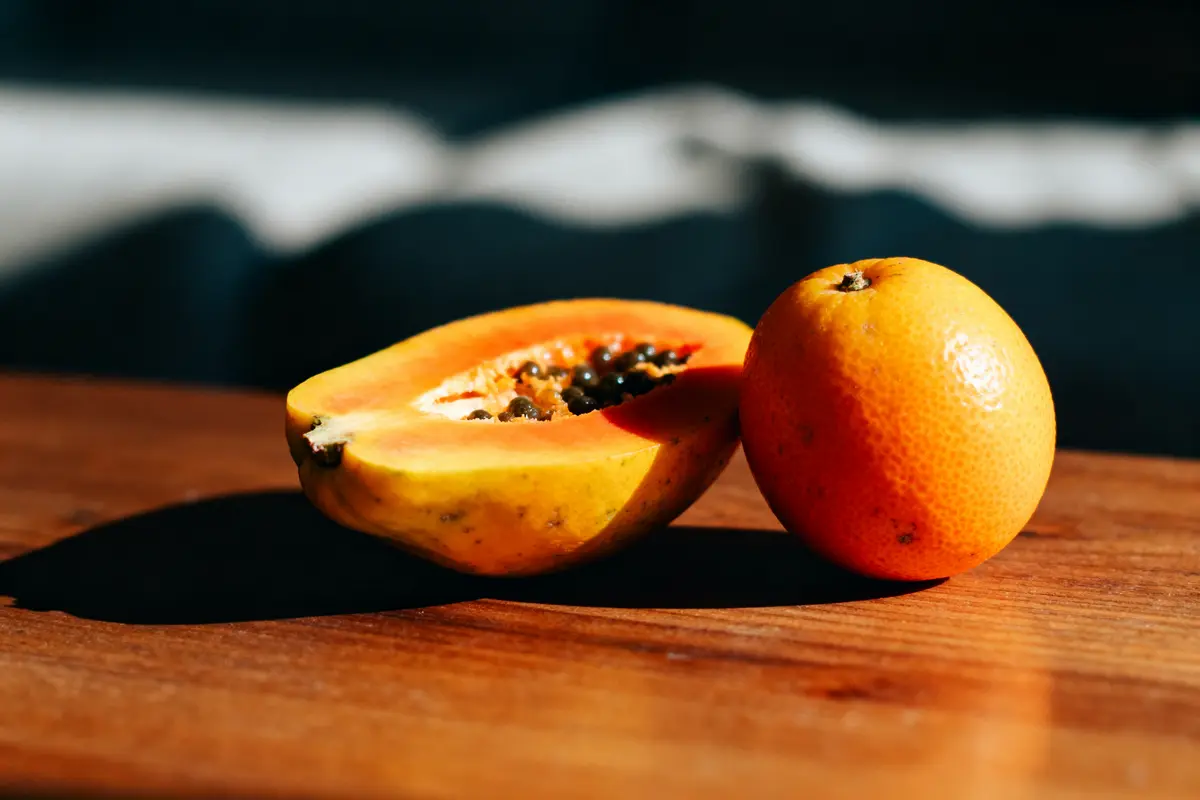 Papaye coupée et orange entière sur table en bois avec éclairage ensoleillé.
