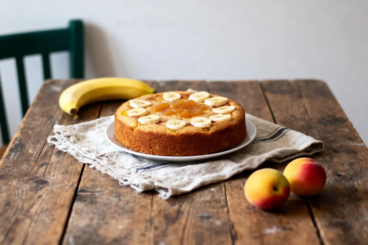Gâteau aux bananes et pêches sur table en bois, avec fruits à côté.